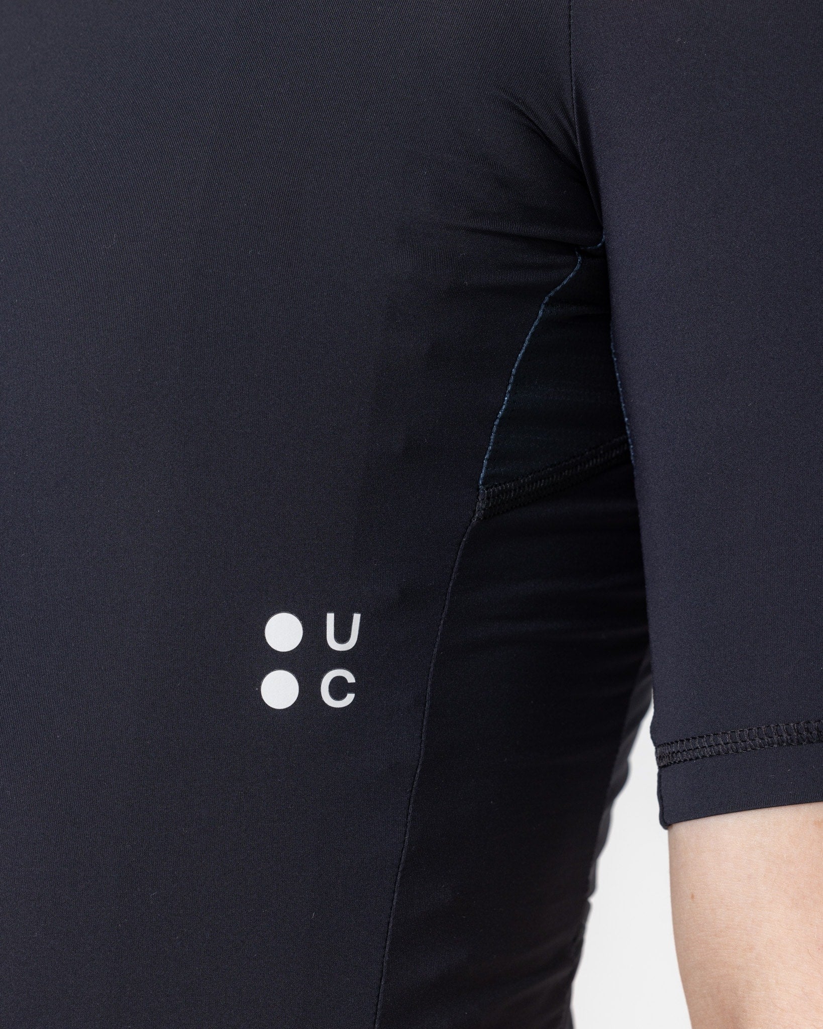 Side panel close-up of the Black Mono Short Sleeve Jersey: white UC dot-mark logo above a breathable mesh underarm.