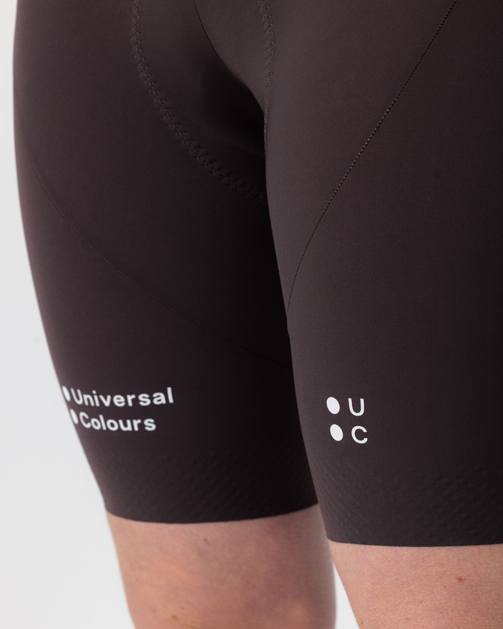 Front leg close-up: tonal Universal Colours leg logos and textured gripper hem on the Heavy Bark Brown Chroma Bib Shorts.