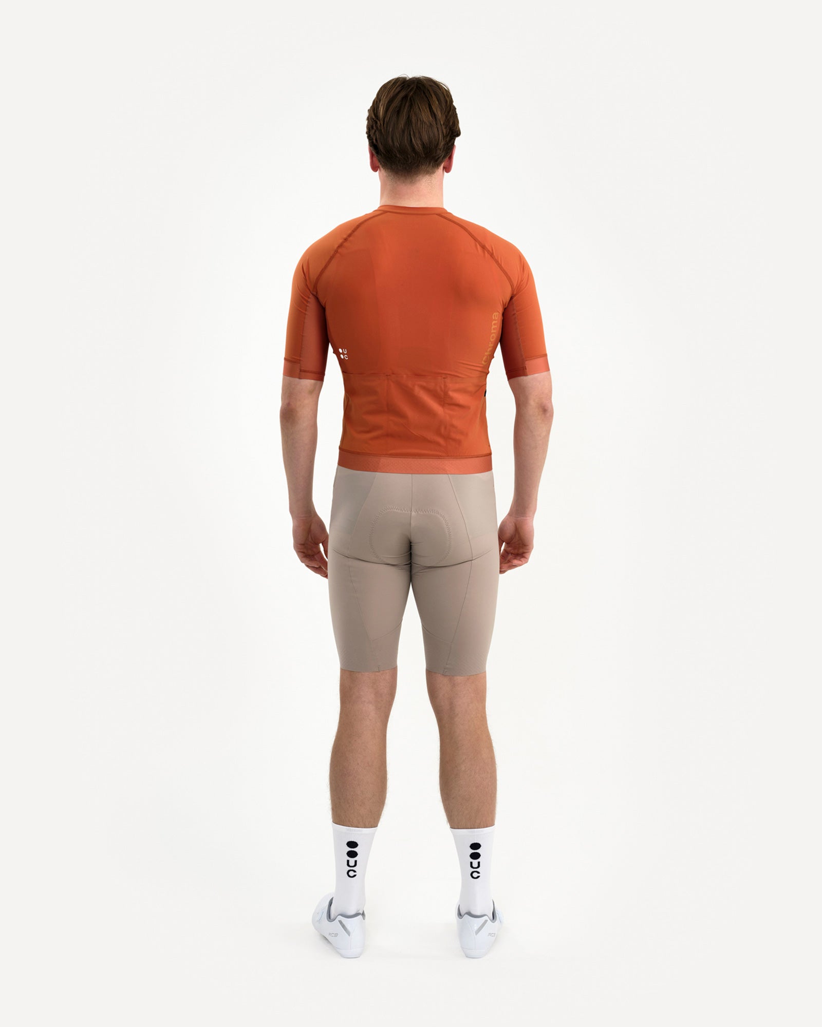 Back view: model in the Men's Chroma Short Sleeve Jersey in Cyprus Copper, paired with stone bib shorts and white UC socks.