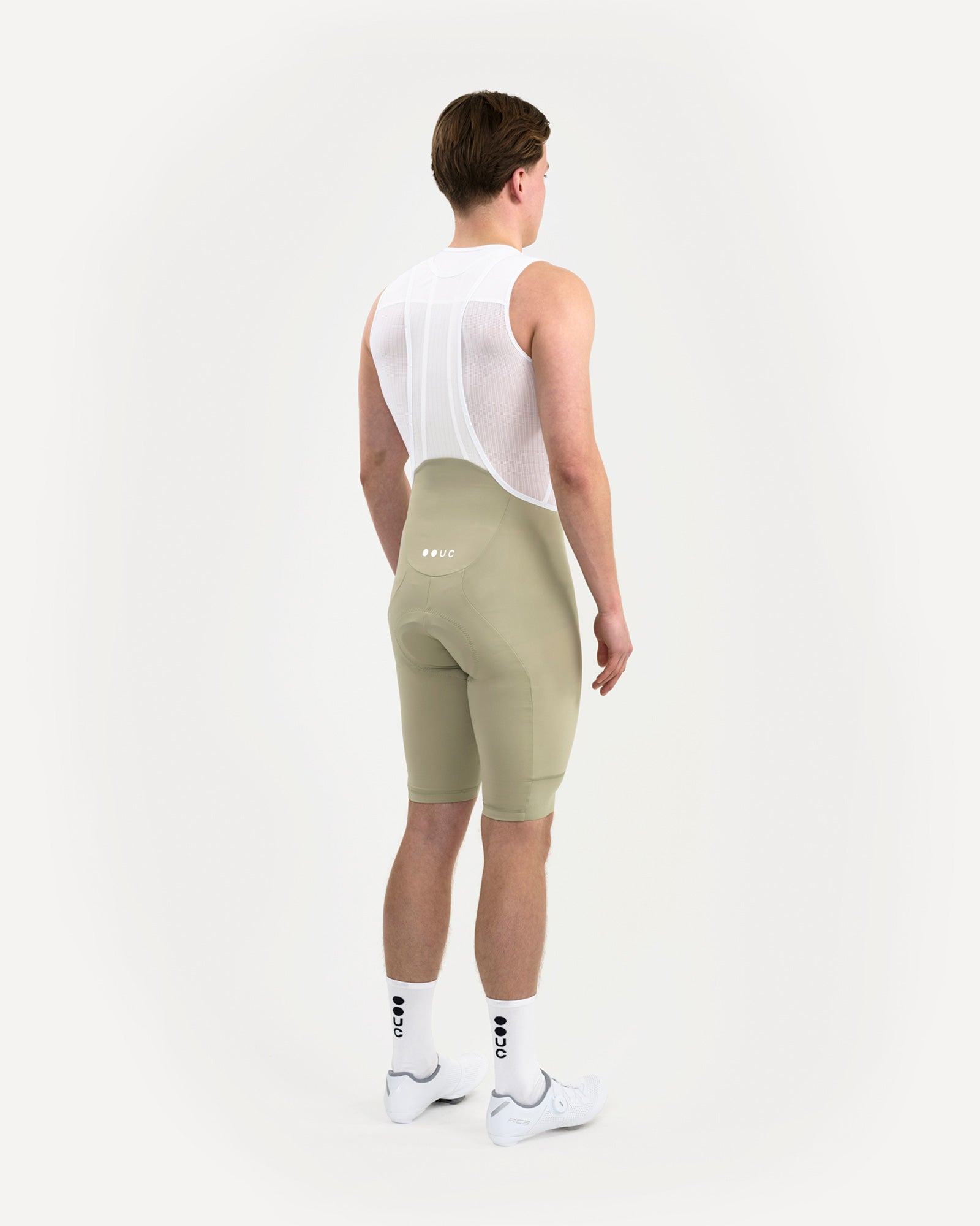 Three-quarter back view of the Men's Mono Bib Shorts 2.0 in Green Tea, with white UC base layer, socks and cycling shoes.