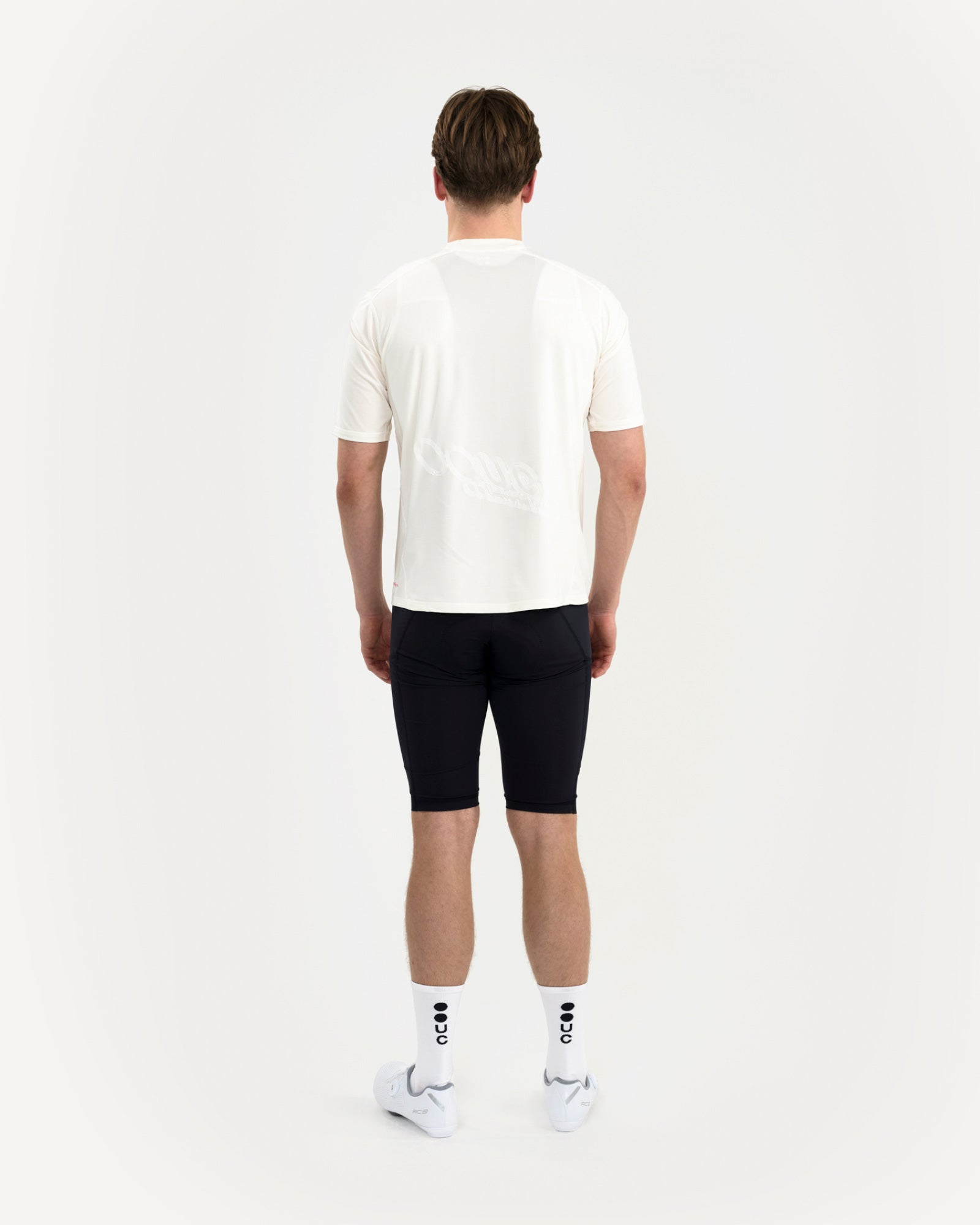 Men's back view: Tech Tee in Off White with tonal script print, black cycling bib shorts and UC-logo white socks.