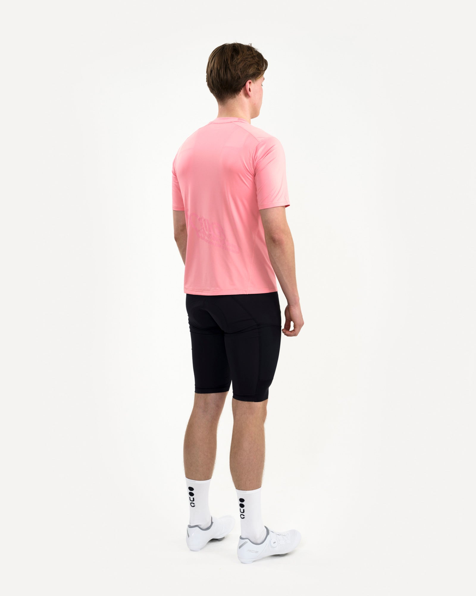 Three-quarter rear view of a male model in the Tech Tee in Washed Pink, paired with black cycling bib shorts.