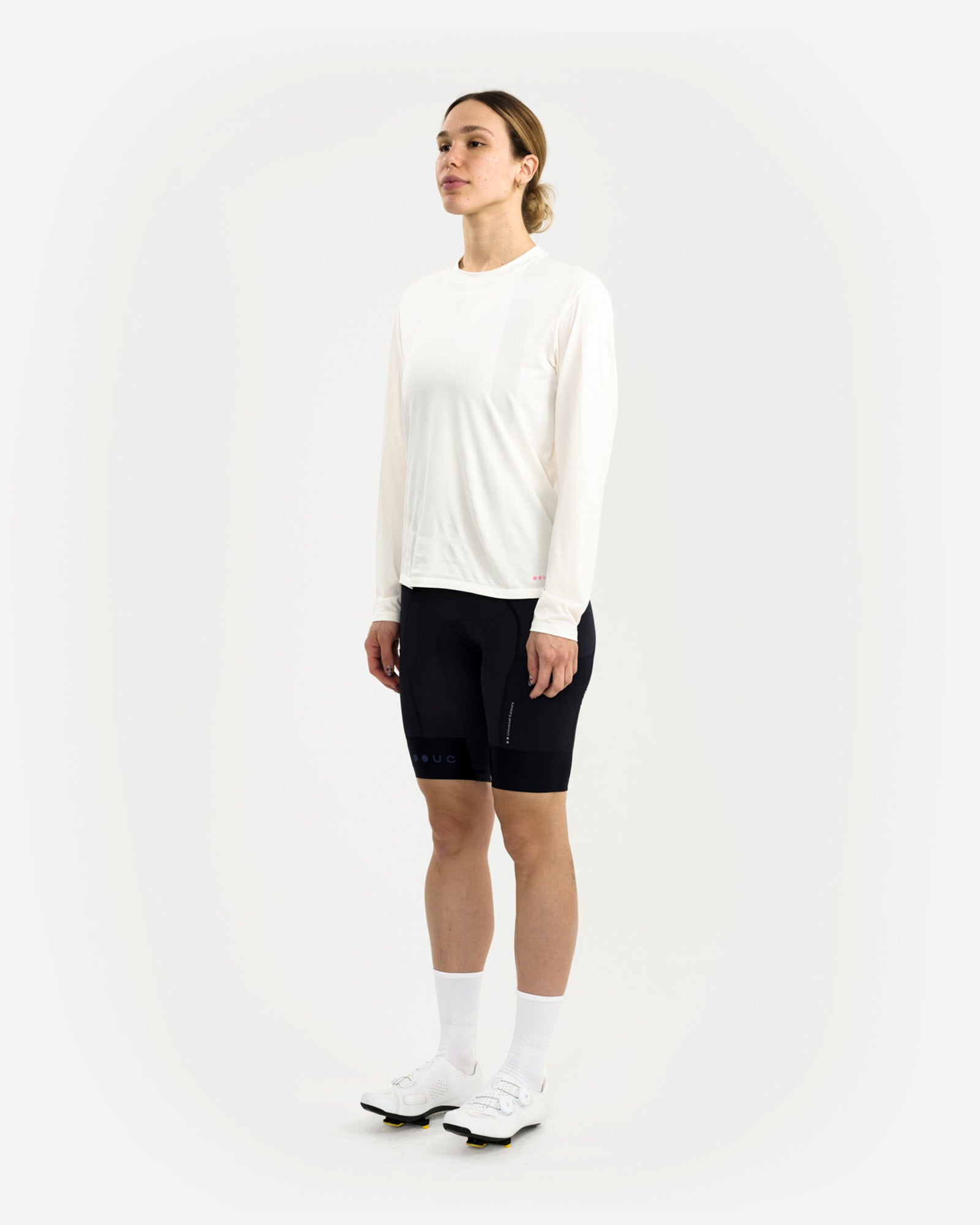 Three-quarter front view: female model in the Long Sleeve Tech Tee in Off White, pink UC hem logo and black bib shorts.