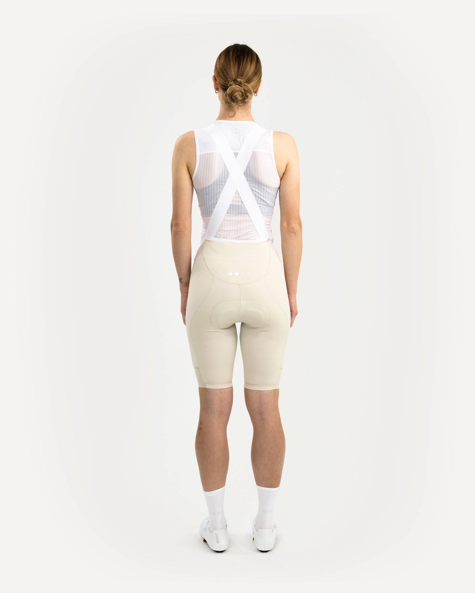 Back view: model in Women's Mono Bib Shorts 2.0 in Off White, paired with a white UC mesh base layer, socks and shoes.