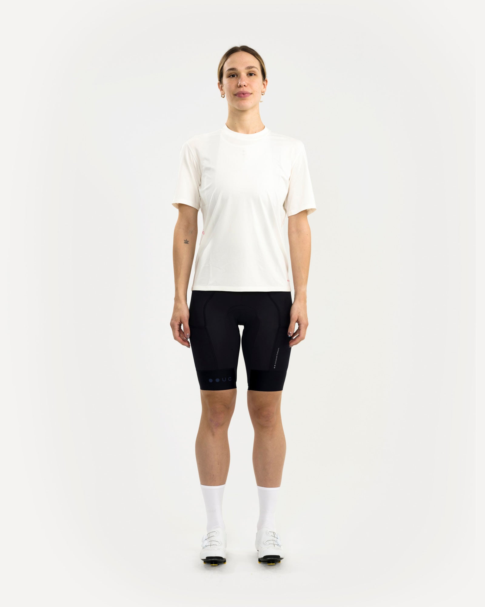 Front view: woman wearing the Tech Tee in Off White with black cycling bib shorts and white road cycling shoes.