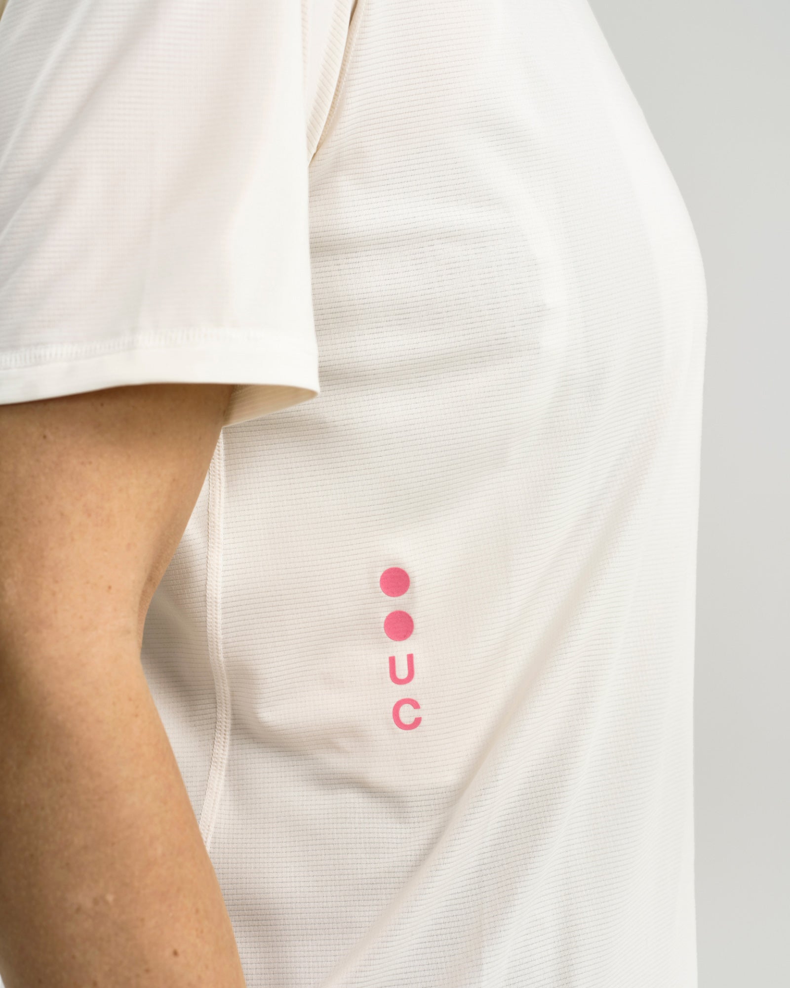 Close-up of the pink UC vertical logo on the front of the Tech Tee in Off White, with fine mesh-knit fabric texture.