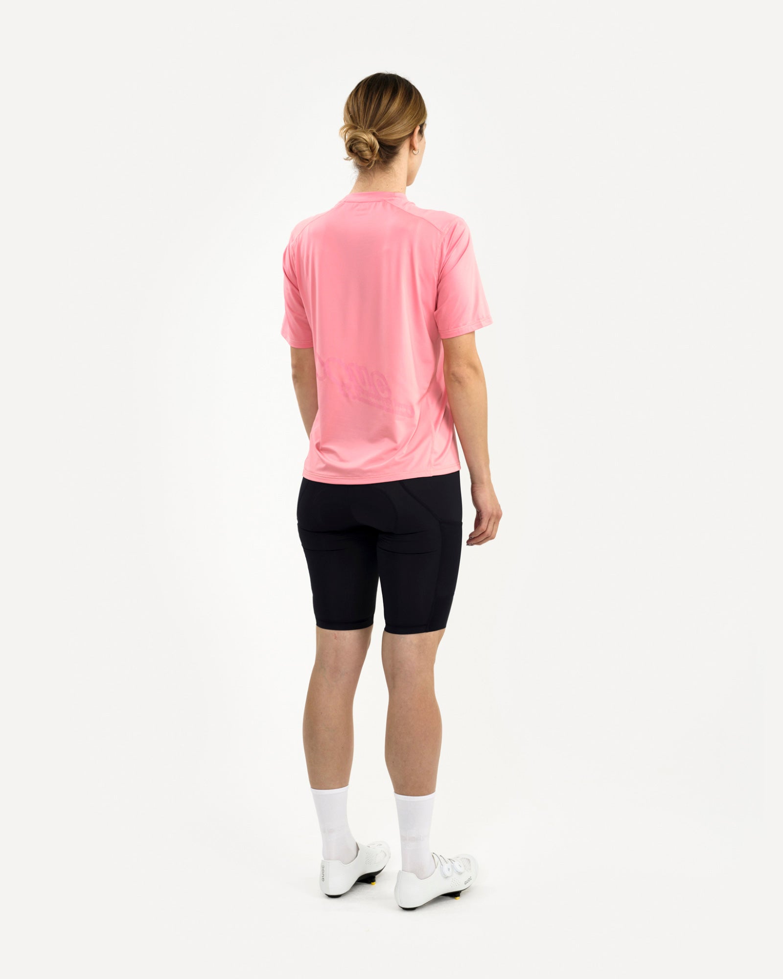 Three-quarter rear view of a female model in the Tech Tee in Washed Pink, paired with black cycling bib shorts.