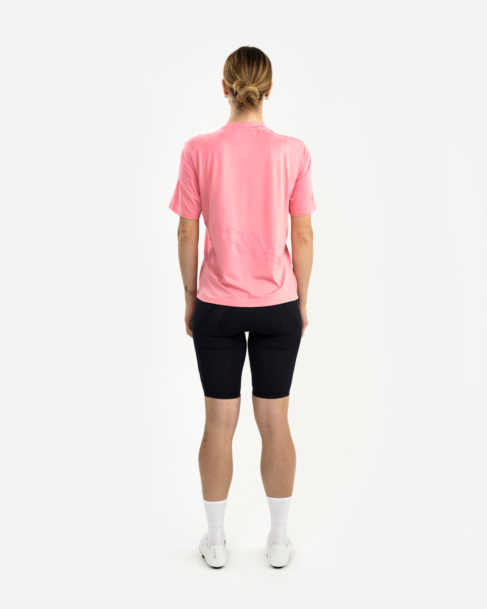 Back view: female model in the Tech Tee in Washed Pink with black bib shorts and tonal cursive back print visible.