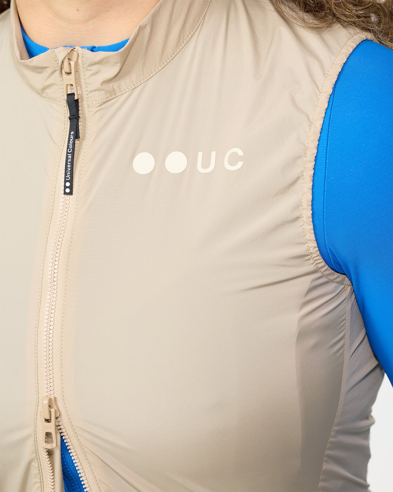 Close-up of the tonal Rice Beige UC chest logo and zip pull on the Women's Mono Gilet 2.0, with cobalt base layer beneath.