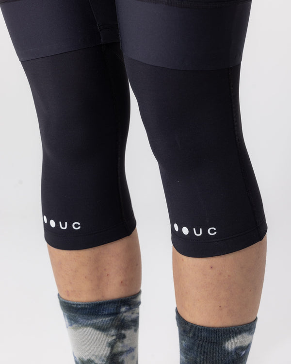 Detail on model: Knee Warmers in Black worn under black bib shorts, with UC roundel and wordmark above the cuff.