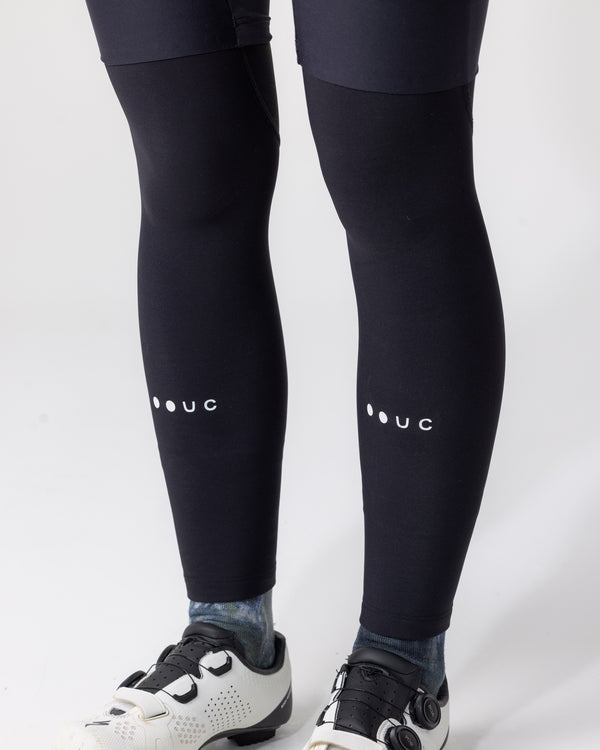 Model wearing the UC Leg Warmers in Black, knees-to-ankles view, pulled over bib tights with white UC wordmark.
