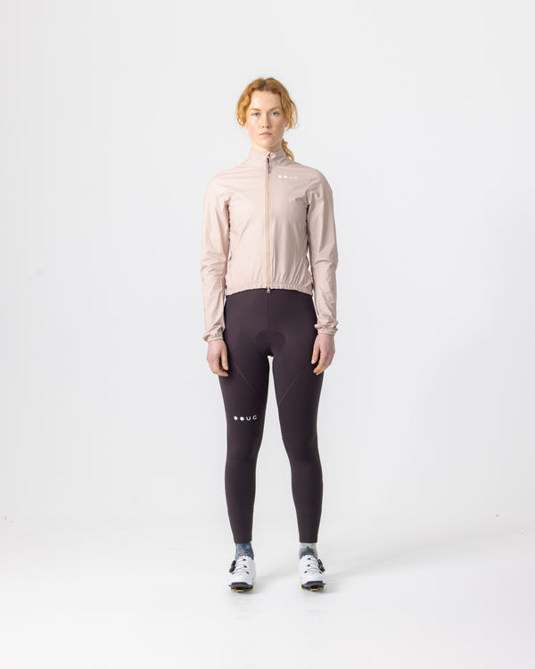 Model wearing the Women's Mono Rain Jacket in Almond Beige, front view, paired with aubergine UC cycling bib tights.