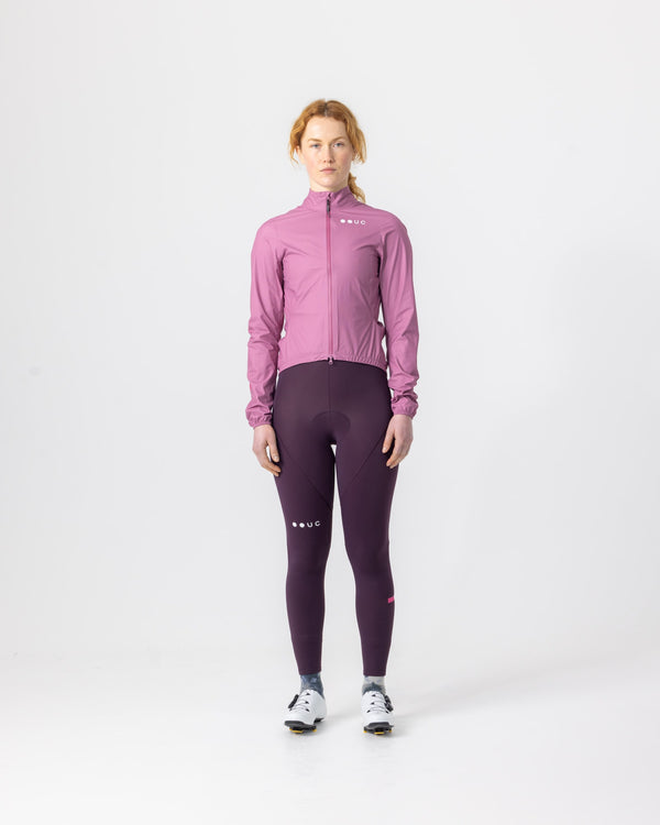 Model wearing the Women's Mono Rain Jacket in Soft Pink, front view, paired with deep aubergine cycling bib tights.
