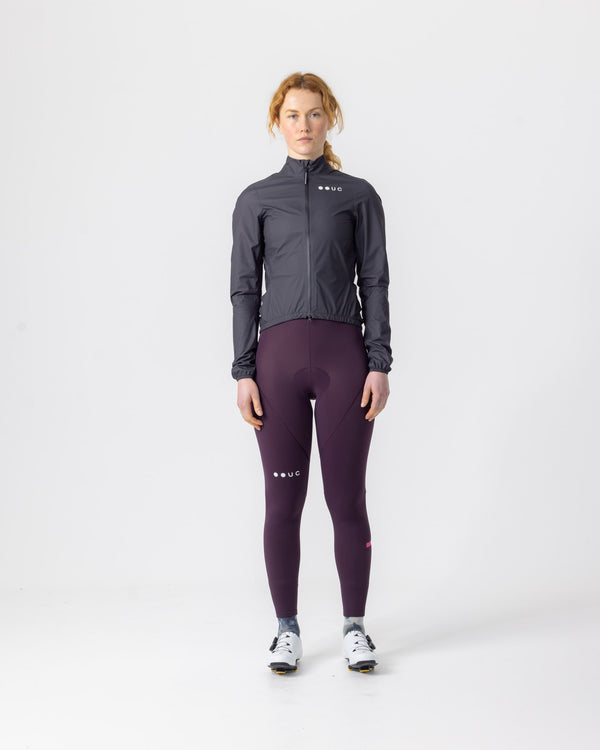 Model wearing the Women's Mono Rain Jacket in Black, front view, paired with aubergine UC cycling bib tights.