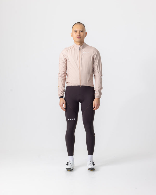 Model wearing the Men's Mono Rain Jacket in Almond Beige, paired with dark Mono bib tights and white cycling shoes.