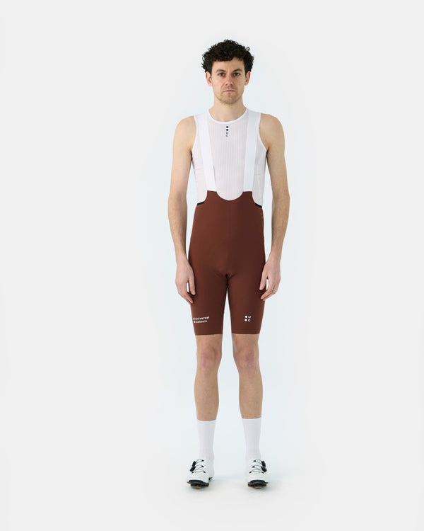 Model wearing the Men's Chroma Bib Short in Brick Red, front view, paired with a white mesh base layer and cycling shoes.