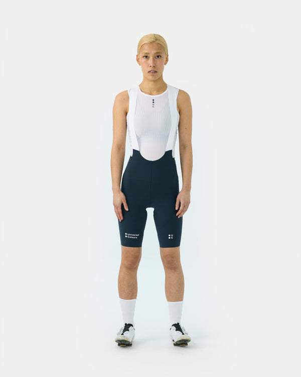 Front view: model wearing the Women's Chroma Bib Short in Navy Blue with a white UC mesh base layer top.
