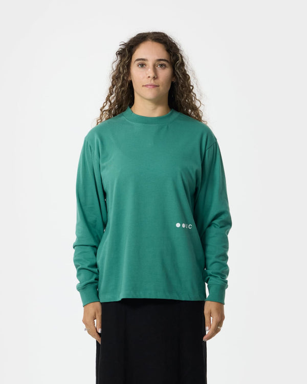 Female model wearing the Unisex Long Sleeve Tee in Bottle Green styled with a black skirt, showing the relaxed boxy fit.