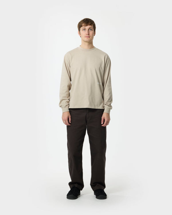 Male model wearing the Unisex Long Sleeve Tee in Oatmeal, styled with dark brown trousers and black trainers.