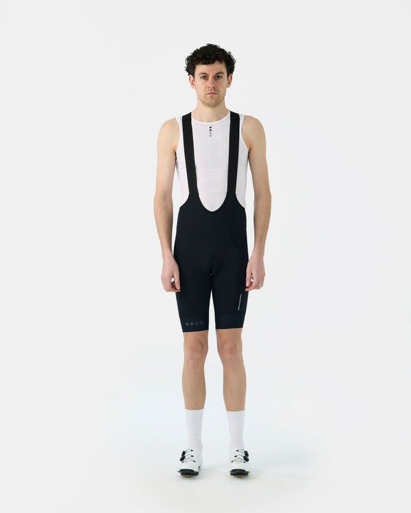 Model wearing the Men's Mono Bib Short 2.0 in Black with a white UC base layer, white socks and white cycling shoes.