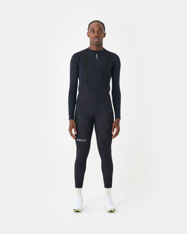 Model wearing the Men's Mono Cargo Bib Tight in Black, paired with a black UC long-sleeve base layer and white cycling shoes.