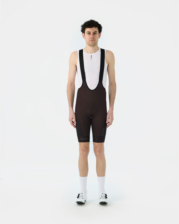 Model wearing the Men's Mono Cargo Bib Shorts in Heavy Bark Brown, front view, with a white UC base layer and cycling shoes.