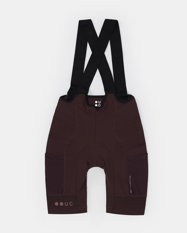 Flatlay of the Women's Mono Cargo Bib Shorts in Heavy Bark Brown: black bib straps, tonal UC chest logo, side cargo pockets.