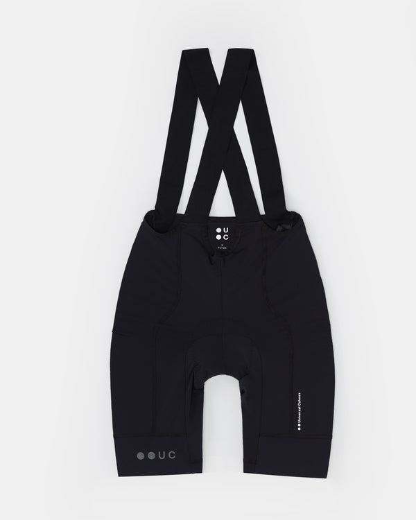 Flatlay of the Women's Mono Bib Shorts 2.0 in Black with crossed bib straps, tonal UC hem logo and chamois seam visible.