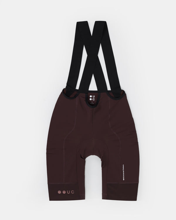 Flatlay of the Women's Mono Bib Shorts 2.0 in Heavy Bark Brown, black crossover straps and tonal UC waistband logo.