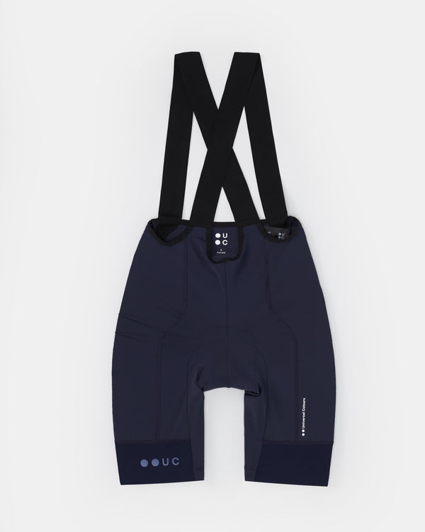 Women's Mono Bib Shorts 2.0 in Navy, flatlay back view: crossed black bib straps, UC dot logo and tonal hem print.