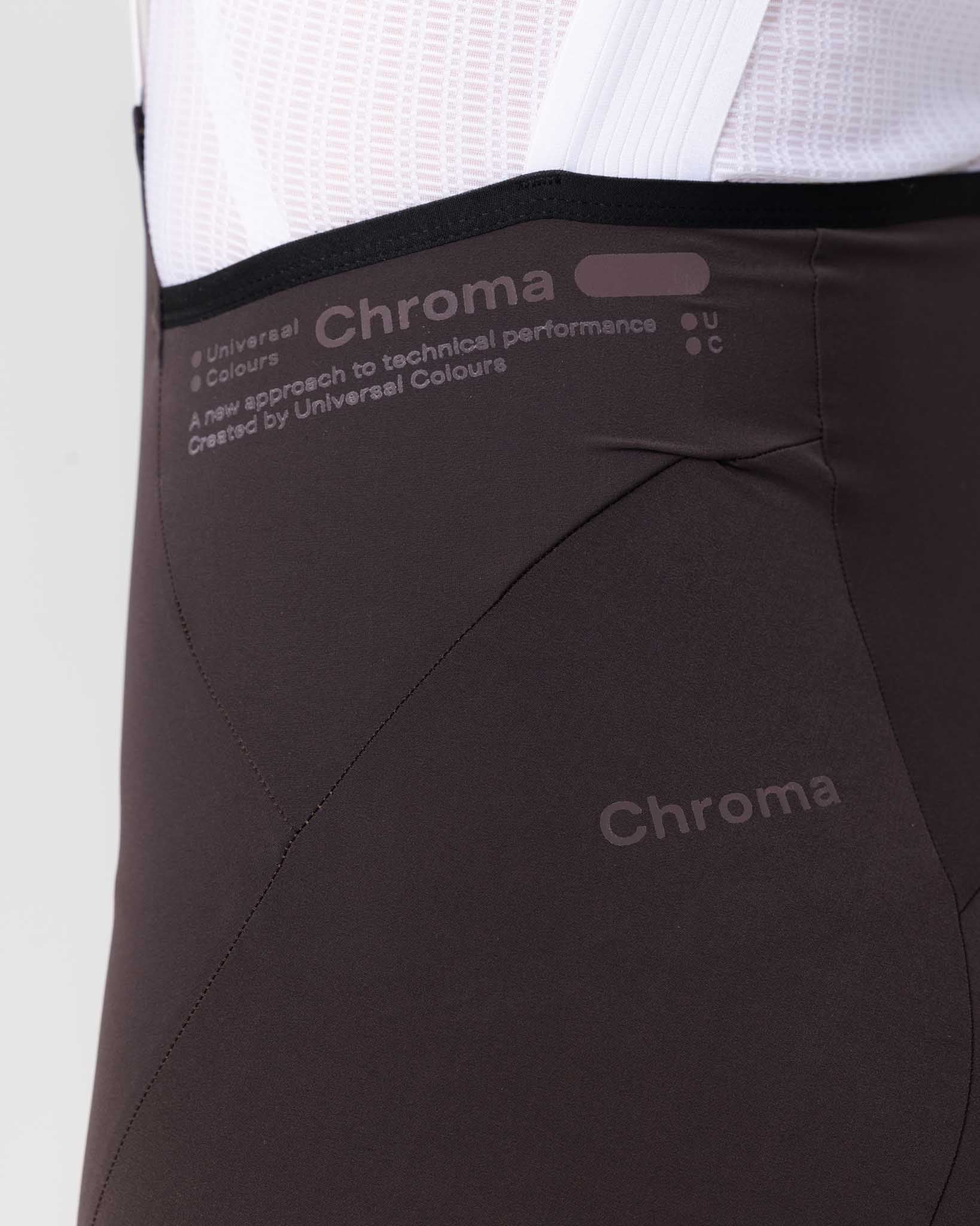 Front waistband close-up: tonal Chroma wordmark on the Heavy Bark Brown Chroma Bib Shorts, white mesh bib straps above.