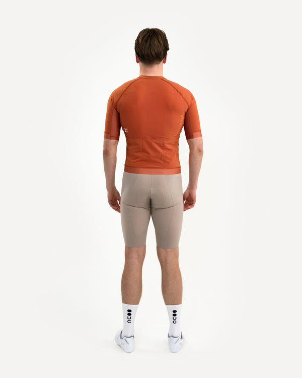 Back view: model in the Men's Chroma Short Sleeve Jersey in Cyprus Copper, paired with stone bib shorts and white UC socks.