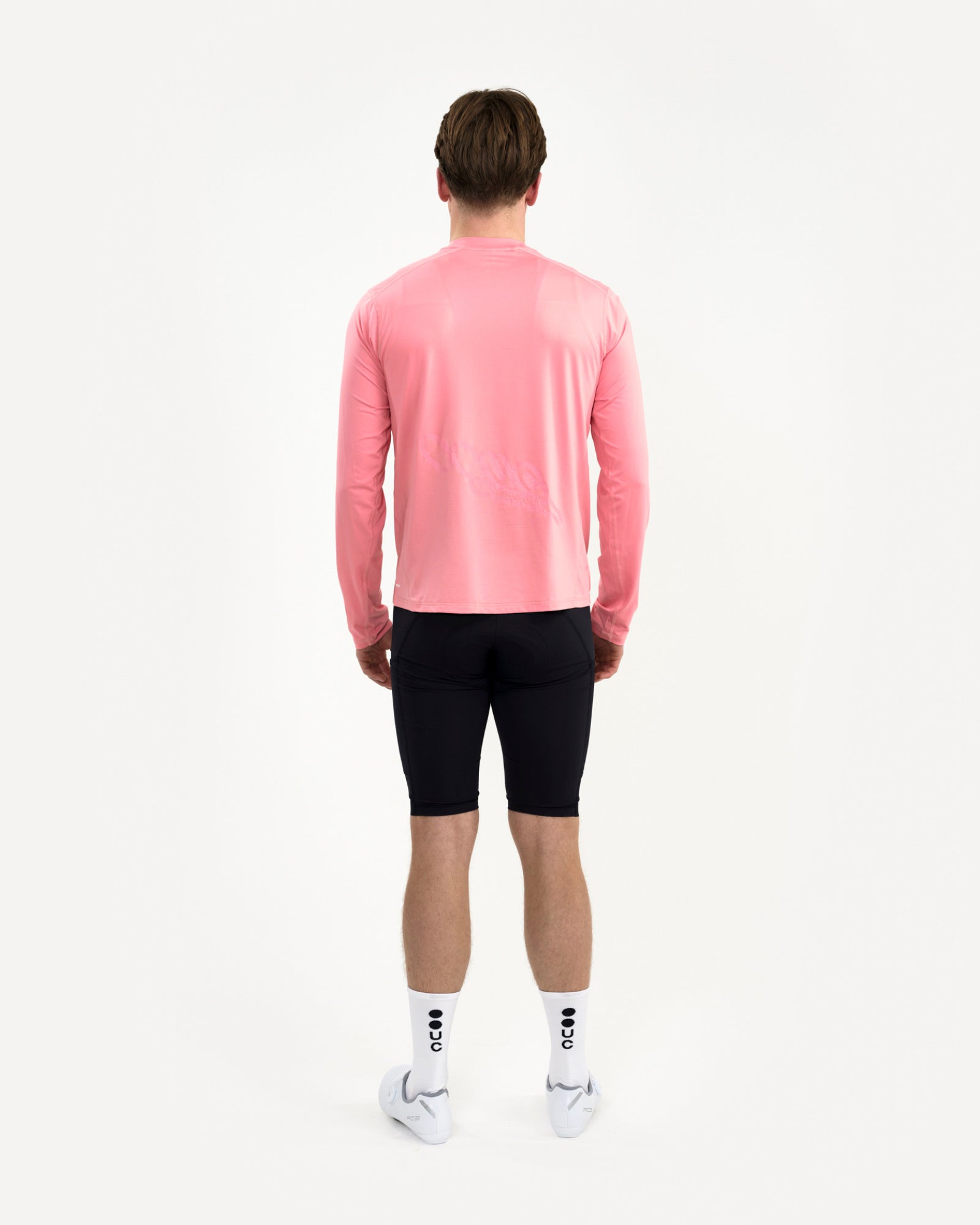 Men's back view of the Long Sleeve Tech Tee in Washed Pink, paired with black bib shorts and UC-branded white cycling socks.