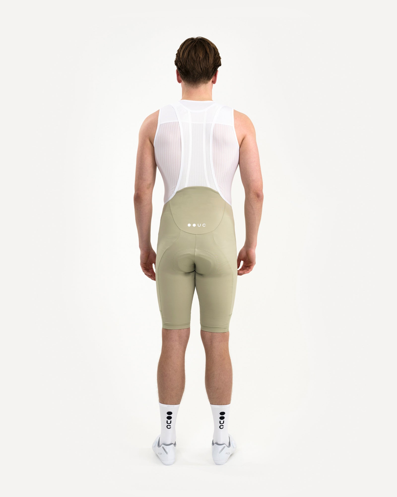 Back view: model in the Men's Mono Bib Shorts 2.0 in Green Tea with white UC mesh base layer, white UC socks and shoes.