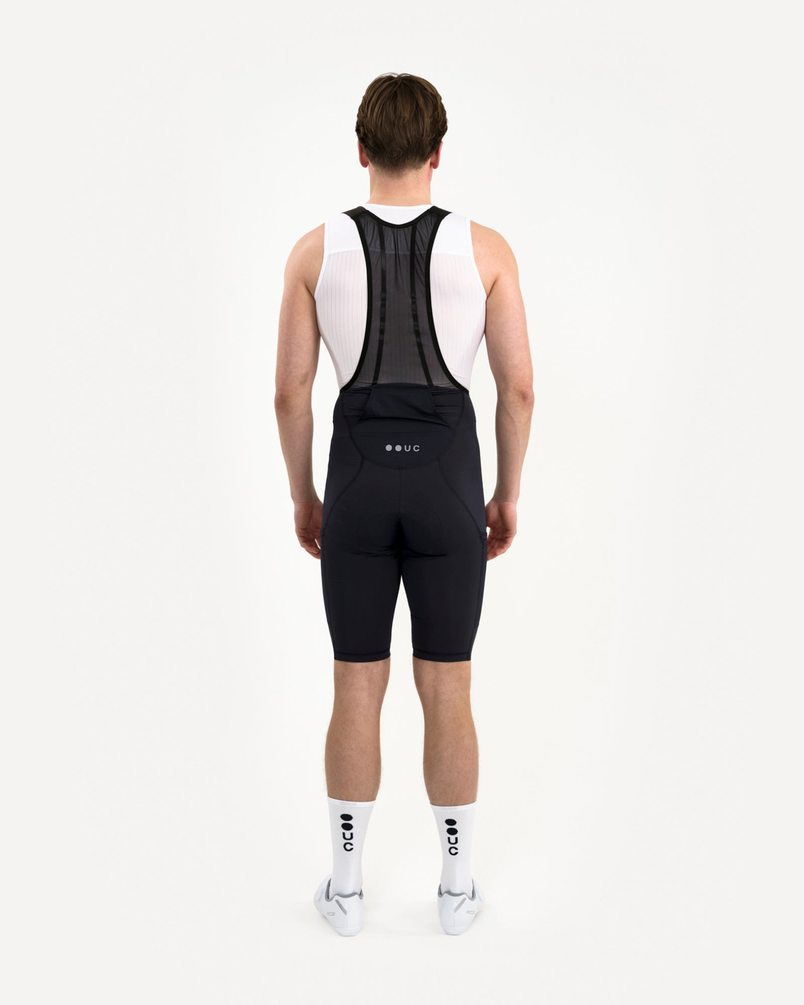 Back view: model in the Men's Mono Cargo Bib Shorts Navy Blue with white UC base layer, dot-logo socks and road shoes.