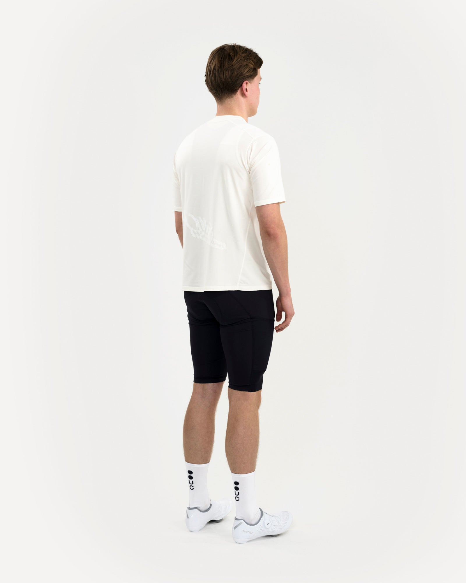 Three-quarter back view: man in the Tech Tee in Off White with black cycling bib shorts and white UC-logo socks.