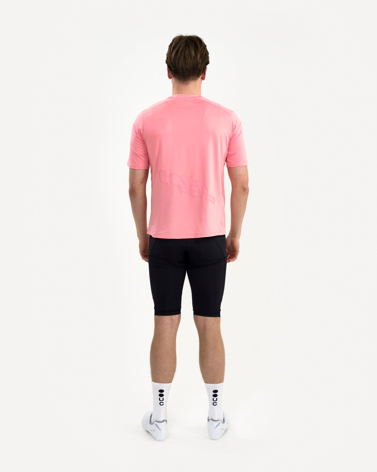 Back view: male model in the Tech Tee in Washed Pink with black bib shorts, white UC socks and white cycling shoes.