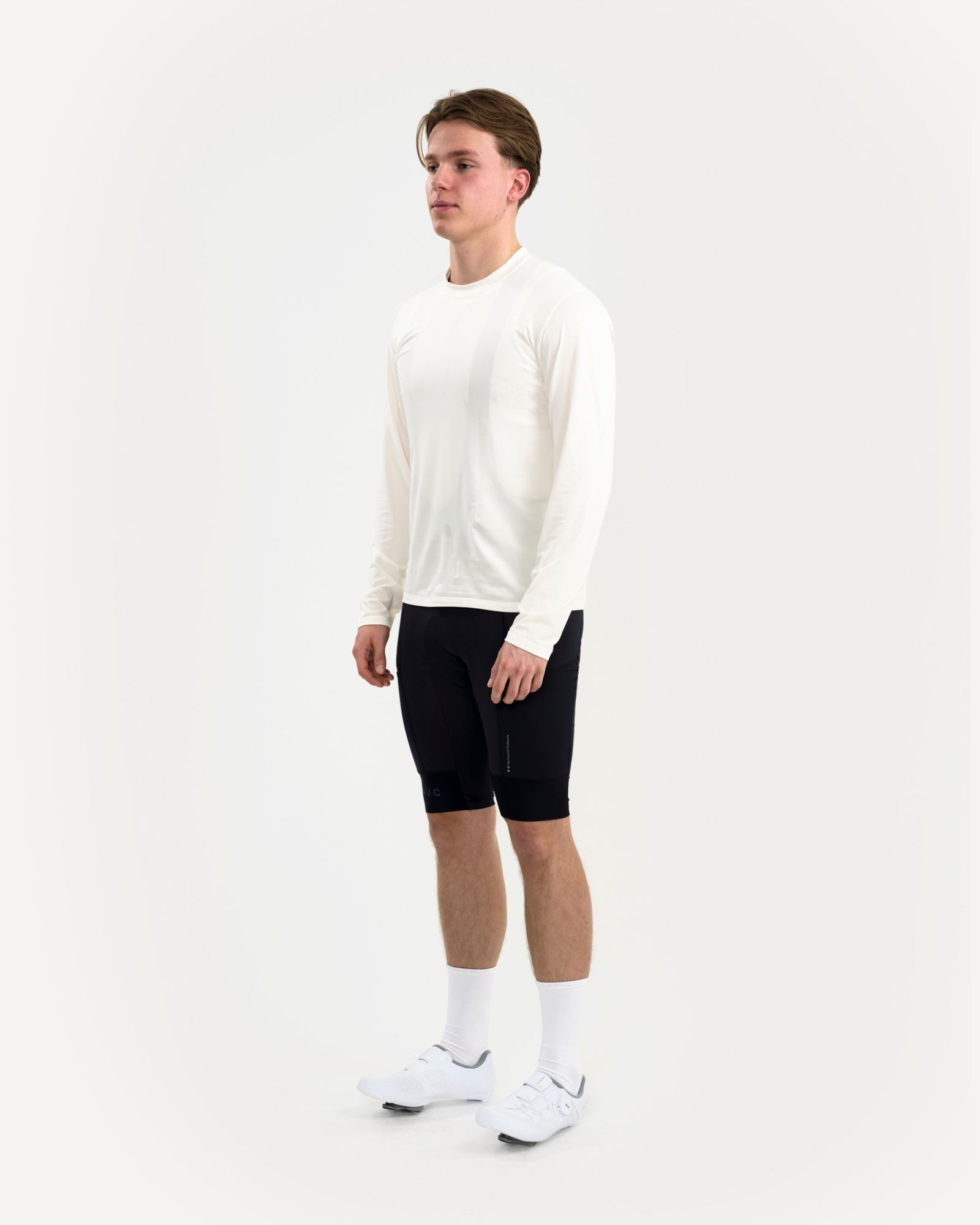 Three-quarter front view: male model in the Long Sleeve Tech Tee in Off White, pink UC hem logo and black bib shorts.
