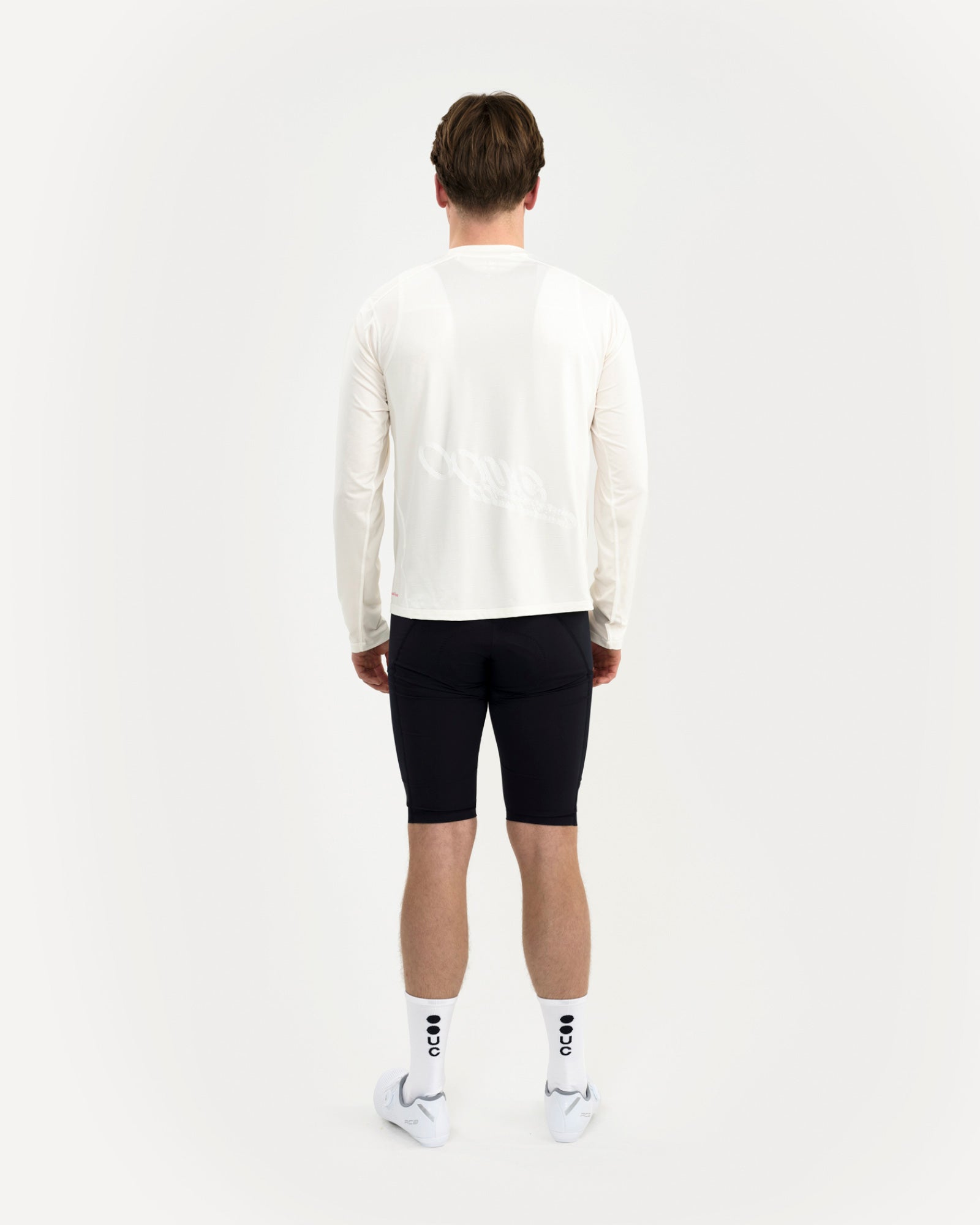Back view: Long Sleeve Tech Tee in Off White on a male model, tonal UC back graphic, with black bib shorts and UC socks.