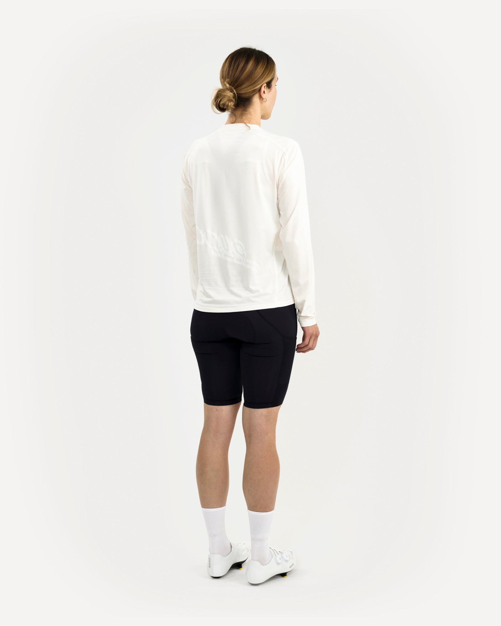 Three-quarter back view of the Long Sleeve Tech Tee in Off White on a female model, tonal UC graphic catching the light.