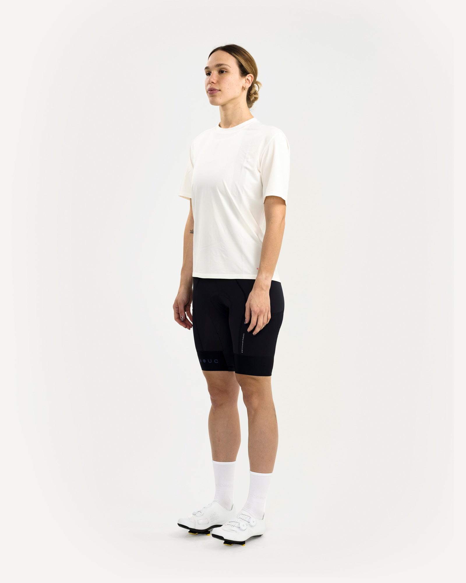 Three-quarter view: woman in the Universal Colours Tech Tee in Off White, paired with black cycling bib shorts.