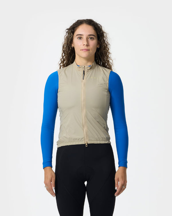 Model wearing the Women's Mono Gilet 2.0 in Rice Beige, zipped over a cobalt blue long-sleeve base layer and black bibs.