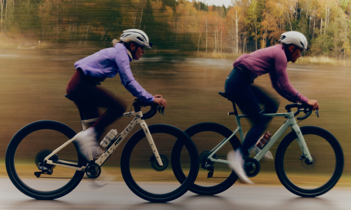 Two riders at speed on a lakeside road with autumn trees behind, one in a lilac jacket and one in a mauve jacket.