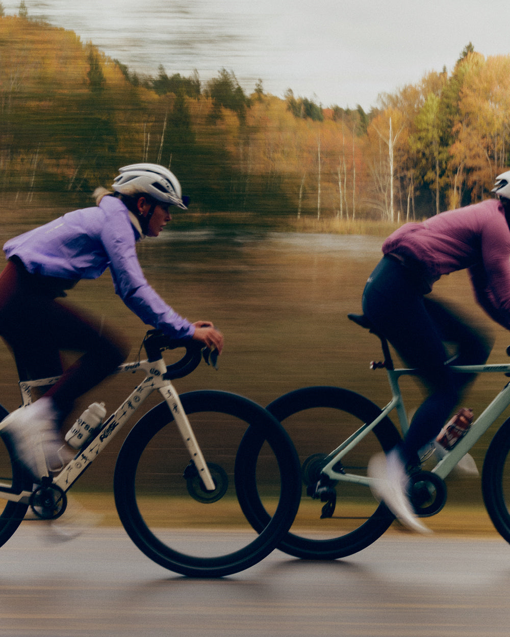 Two riders at speed on a lakeside road with autumn trees behind, one in a lilac jacket.