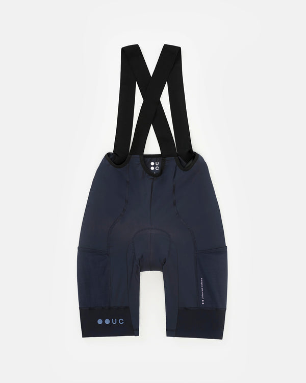Flatlay rear view: Women's Mono Cargo Bib Short in Navy Blue with black X-cross bib straps and tonal UC leg-cuff logo.