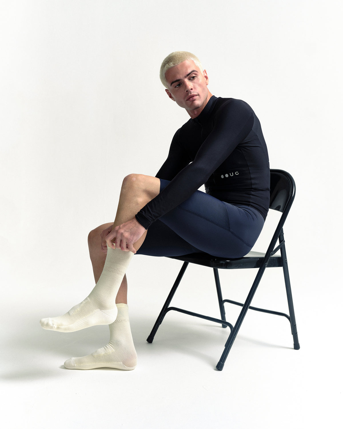 Seated model pulling on the Merino Winter Socks in Off White, full-length side view with navy bib shorts and a black long-sleeve top.